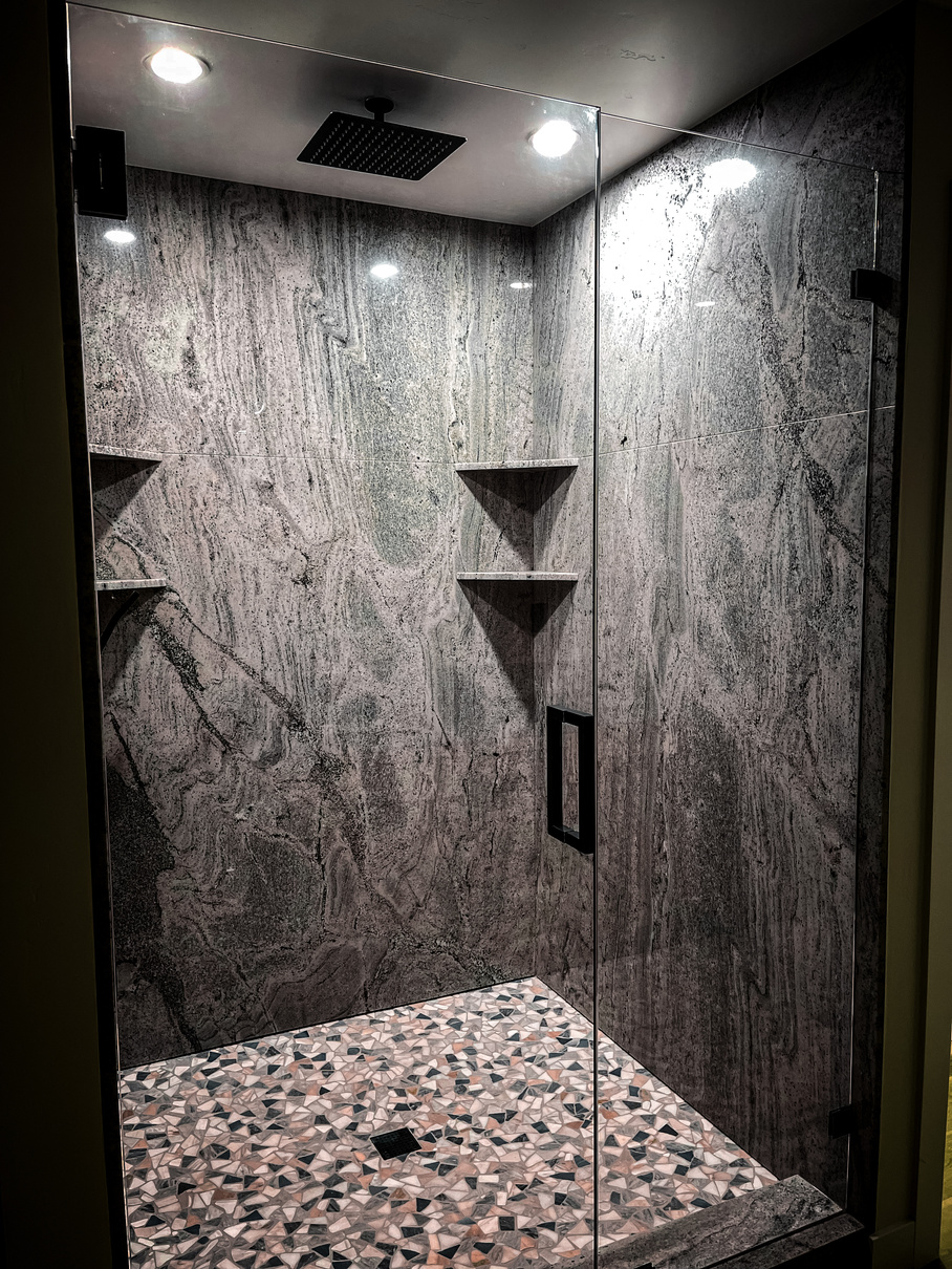 a freshly remodeled walk-in shower with a glass door, waterfall shower head, and grey stone walls with tile floor.